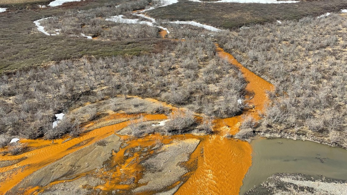  An aerial view of a river with orange-colored water flowing through a snow-covered landscape in Alaska.