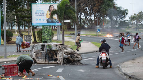 Residents of Noumea in New Caledonia pass by a burned-out car near a looted supermarket in the N'Gea district, May 14, 2024.