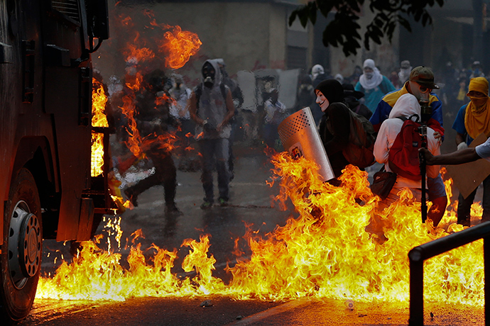 An anti-government protester, wearing a Guy Fawkes mask, stands with a shield near flames from molotov cocktails thrown at a water cannon by anti-government protesters during riots in Caracas April 20, 2014 (Reuters / Jorge Silva)
