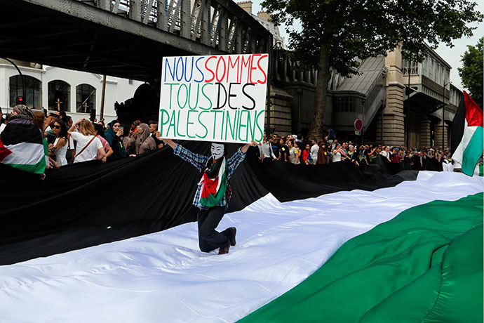 A protester holds a placard reading "We are all Palestinians" as he kneels on a giant Palestinian flag near the Barbes-Rochechouart aerial metro station prior to the departure of a demonstration, banned by French police, in Paris on July 19, 2014 to denounce Israel's military campaign in Gaza and show support for the Palestinian people. (AFP Photo / Francois Guillot)