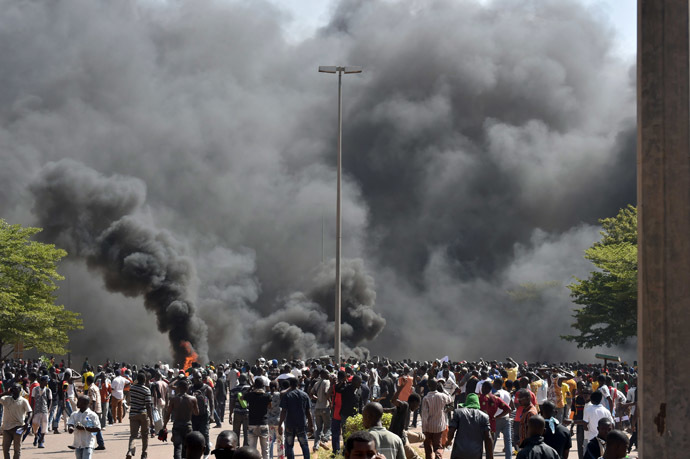 People stand in front of smoke rising from the Burkina Faso's Parliament, where demonstrators set cars on fire parked in a courtyard of the Parliament, on October 30, 2014 in Ouagadougou, as they protest at plans to change the constitution to allow President Blaise Compaore to extend his 27-year rule. (AFP Photo/Issouf Sango)
