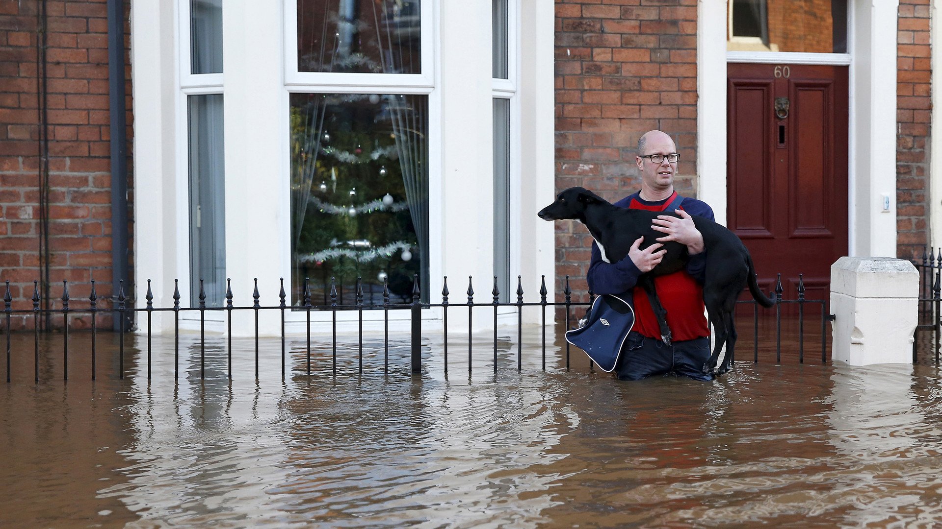 رجل يحمل كلبه أمام بيته على ذراعيه في الشارع الذي غمرته مياه الفيضانات بمدينة كارليل، 6 ديسمبر/كانون الأول