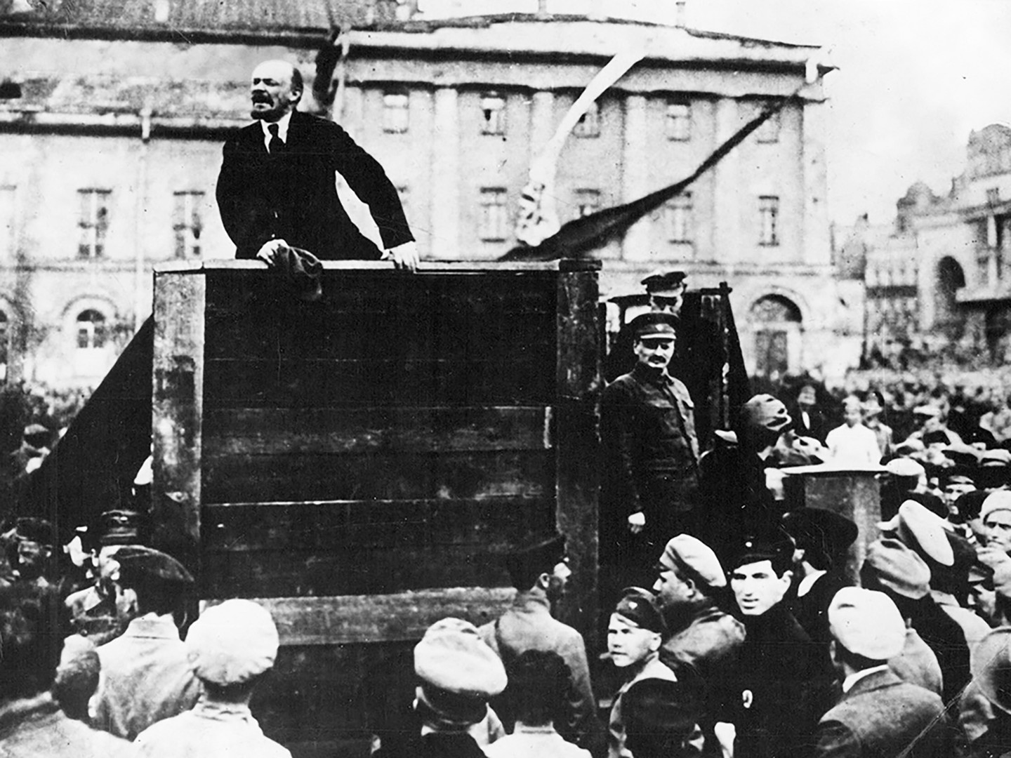 Vladimir Lenin berpidato di tengah kerumunan orang di Lapangan Sverdlov di Petrograd pada 1919. Lev Trotsky berdiri di sebelah kanannya.