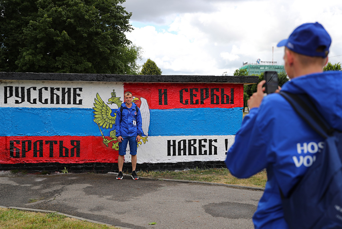 Voluntários da Copa de 2018 posando para fotografia ao lado de mural que mostra as bandeiras da Rússia e da Sérvia e a mensagem “Os russos e os sérvios são irmãos para sempre”, no centro de Kaliningrado