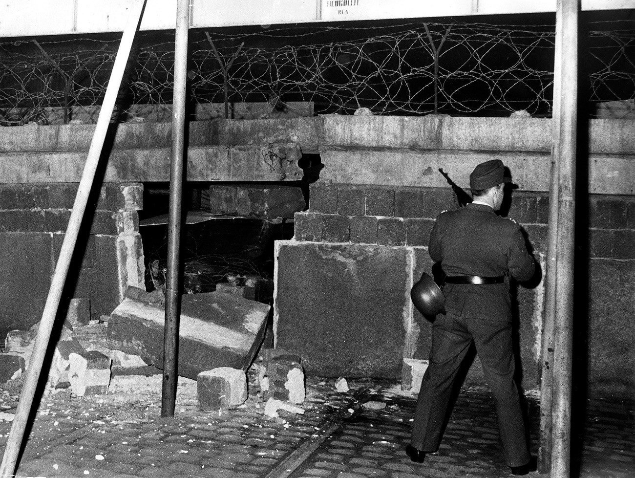 Polisi Jerman Timur menjaga tembok yang hancur ditabrak mobil pengangkut personel lapis baja yang dikendarai Wolfgang Engels dalam upaya pelarian dirinya ke Barat.