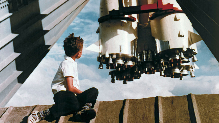 Млад посетител на Изложбата на достигнувањата на народното стопанство на СССР седи на платформа пред ракета „Восток“ близу павилјонот „Космос“.