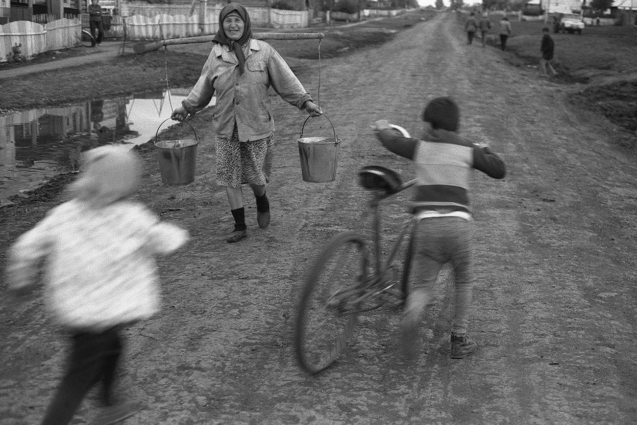 Pemandangan di desa. Seorang anak dengan sepeda dan seorang perempuan tua membawa ember, 1979.