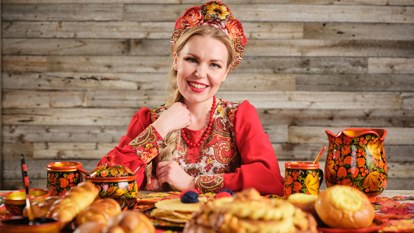 Eine Frau, gekleidet in traditionelle russische Kokoshnik-Kopfbedeckung, sitzt an einem Tisch mit Feiertagsbroten.