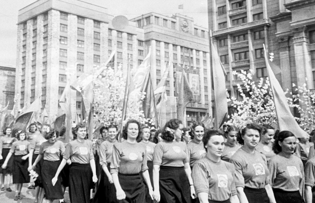 Femmes défilant avec des fleurs et des drapeaux lors de la parade du 1er mai près du bâtiment de la Douma, 1950

