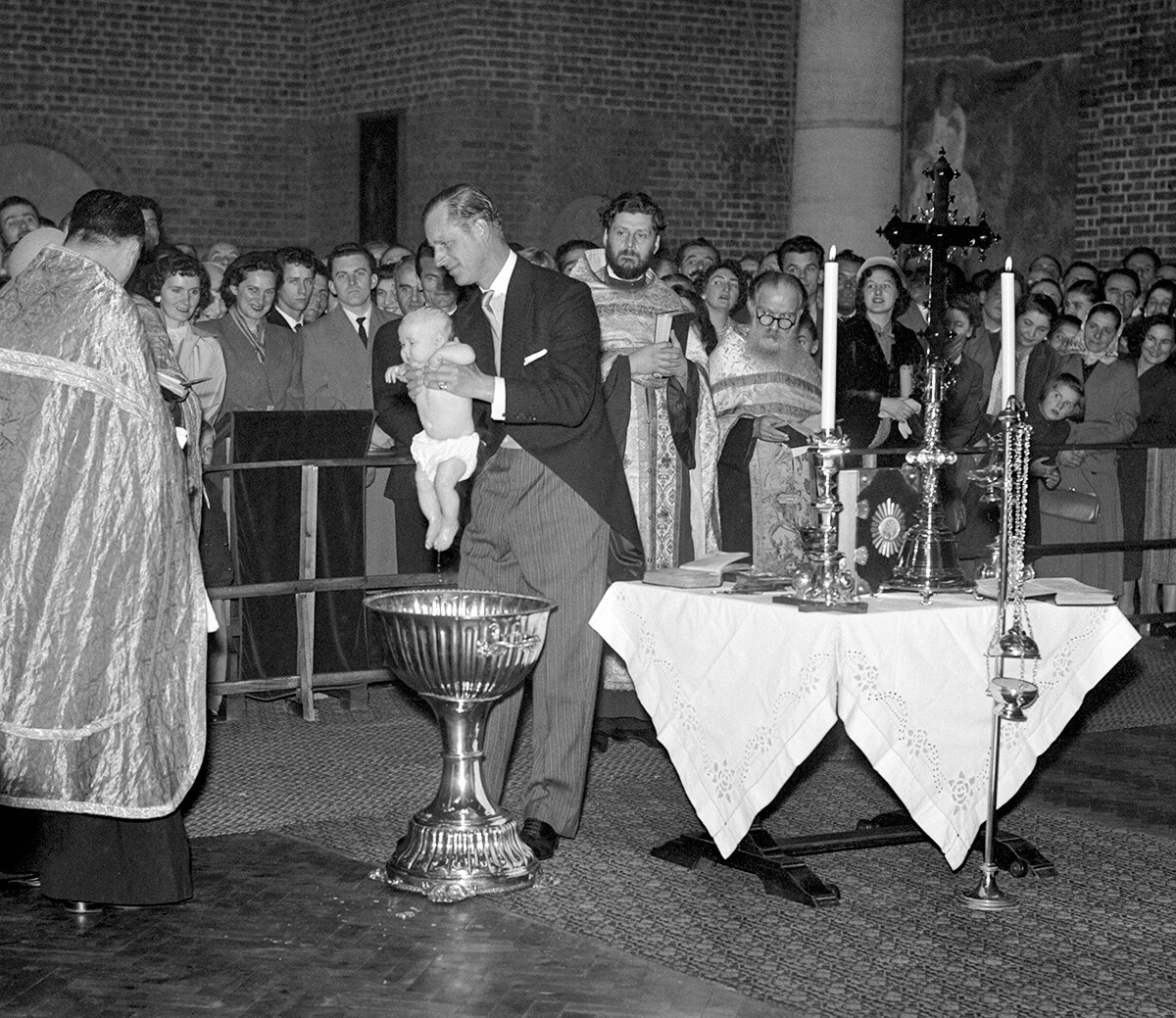 Le duc d'Édimbourg, qui est le parrain de l'enfant, soulève la petite princesse Maria («Tania») des fonts baptismaux après son baptême à l'église orthodoxe serbe. Le bébé est la fille de la nièce du duc, la princesse Christine de Hesse-Cassel, et du prince André de Yougoslavie.