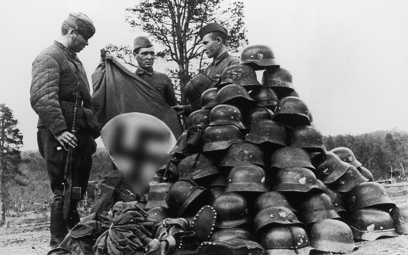 Tres soldados soviéticos muestran una bandera nazi y un montón de cascos y botas militares, capturados tras la destrucción de un regimiento de campo de la Luftwaffe alemana cerca de Múrmansk, en la URSS. 1942
