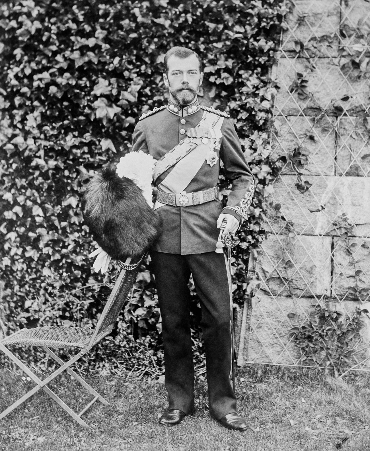  Photograph of Nicholas II, Emperor of Russia standing outside Balmoral. He is wearing British military uniform and is holding a bearskin hat in his right hand and a sword in his left. 