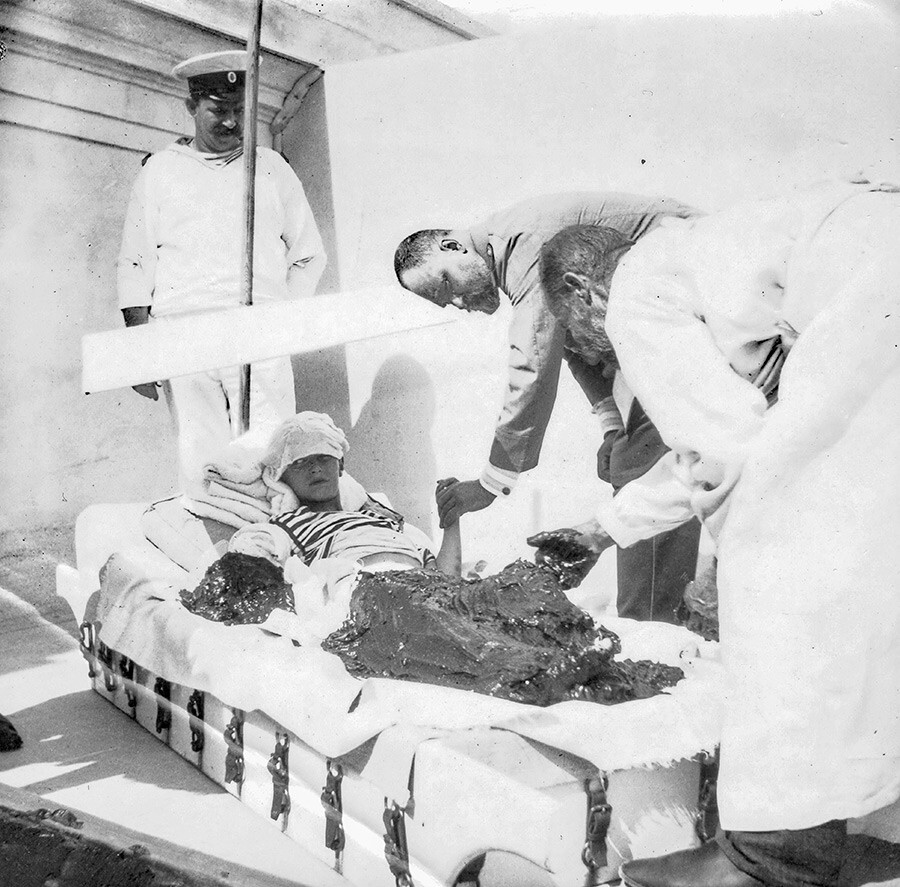 O tsarévitch Alexei durante tratamento com banho de lama para aliviar os sintomas da hemofilia em Livadia, na Crimeia. Ao fundo, em pé, o marinheiro que cuidava dele, Andrêi Derevenko. O doutor Botkin segura a mão esquerda do tsarêvitch.