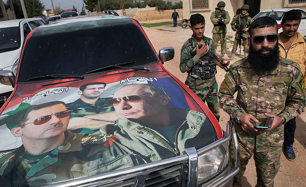 Syrian and Russian solders, who escort a group of journalists in the background, stand near a car covered by collage showing photos of faces of Russian President Vladimir Putin, Syrian President Bashar Assad and a Syrian general, President's Assad brother, Maher Assad, center, in Maarzaf, about 15 kilometers west of Hama, Syria, March 2, 2016.