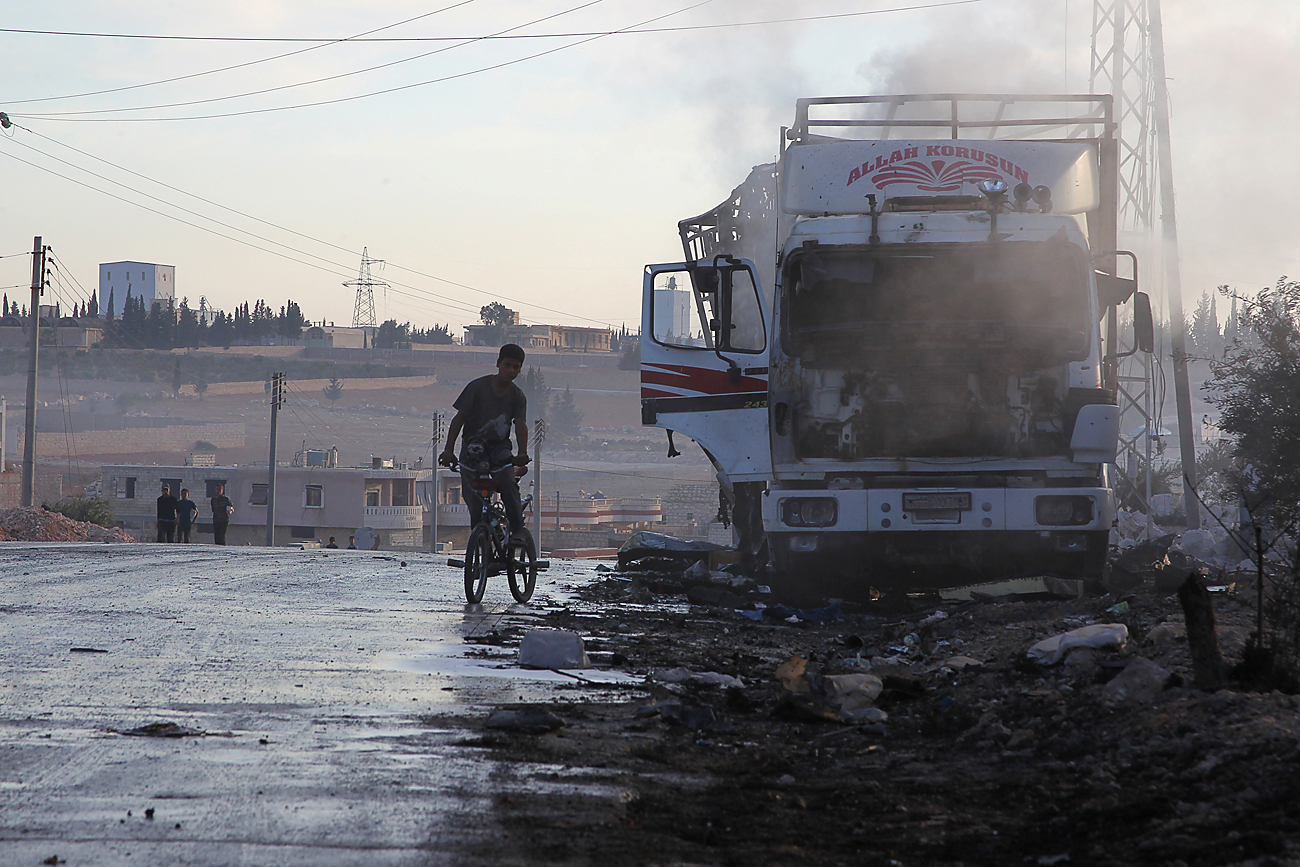 Menino de bicicleta observa caminhão de ajuda humanitária destruído.