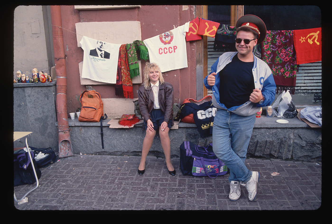 A woman and man in western style clothing near a collection of Soviet and Russian souvenirs on Arbat street, Moscow in 1991. Souvenir items include t-shirts displaying CCCP and photo of Mikhail Gorbachev. 