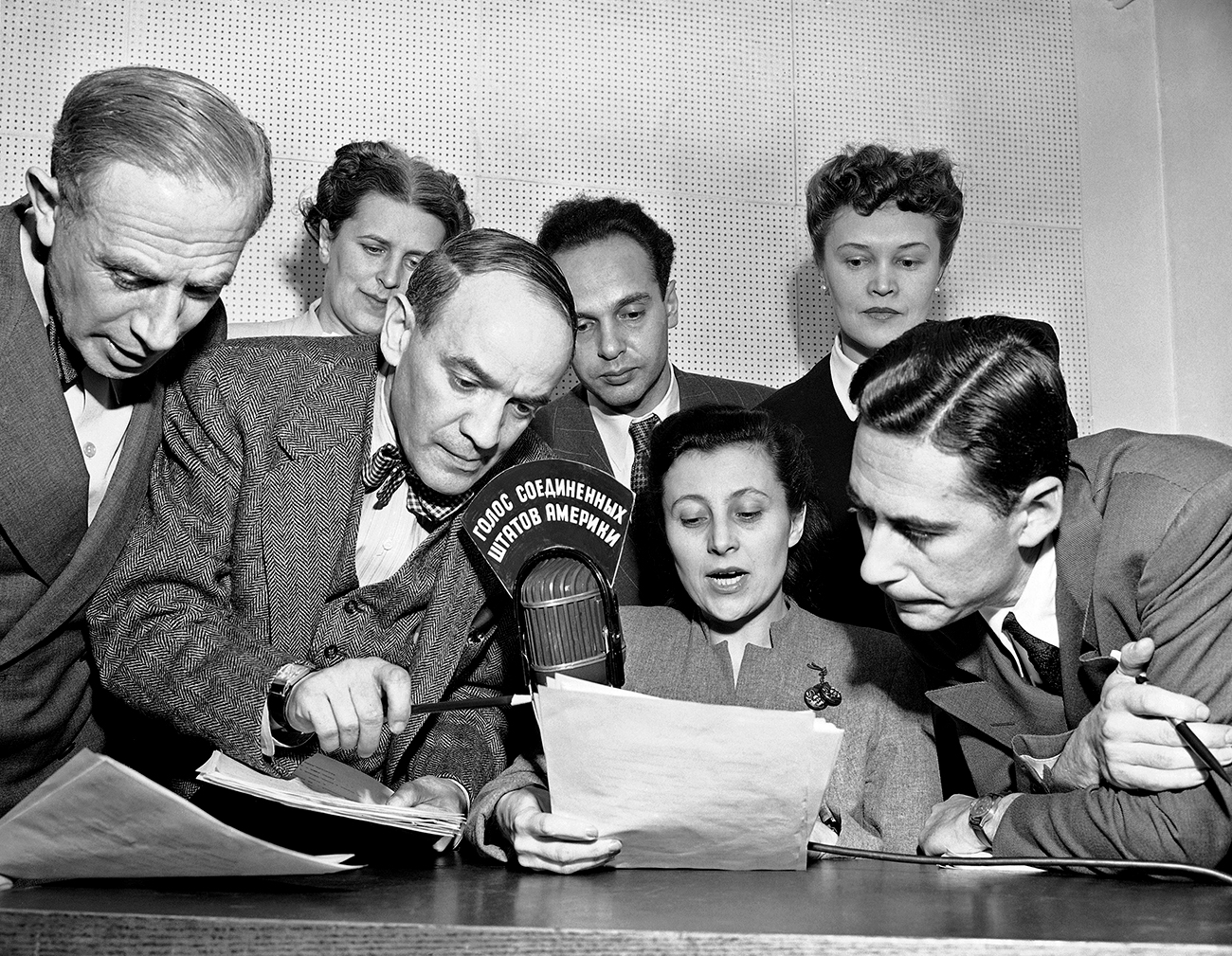 A group of State Department announcers huddle around the microphone after the initial shortwave broadcast in Russian to Russia from New York City on Feb. 17, 1947. The announcers, Russian-born American citizens, are (front row) Boris Brodenov; James Schigorin; Elena Bates (seated); Victor Franzusoff; (rear row) Katherine Elene; Vladimir Postman and Tatiana Hecker. Sign in Russian on the microphone means "The Voice of the United States of America."