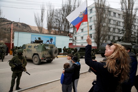 A woman waves a Russian flag as armed servicemen wait near Russian army vehicles outside a Ukrainian border guard post in the Crimean town of Balaclava March 1, 2014. Source: Reuters