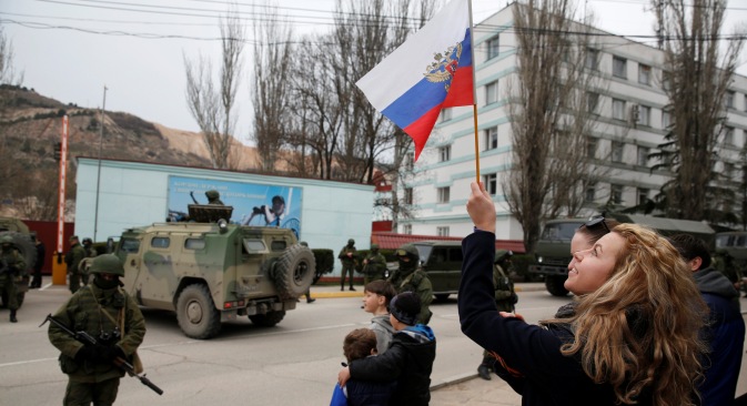 A woman waves a Russian flag as armed servicemen wait near Russian army vehicles outside a Ukrainian border guard post in the Crimean town of Balaclava March 1, 2014. Source: Reuters