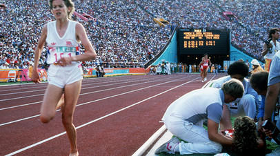 Зола Бадд преодолевает очередной круг финала бега на 3000 метров на Олимпиаде в Лос-Анджелесе, пока врачи и тренеры оказывают помощь Мэри Деккер